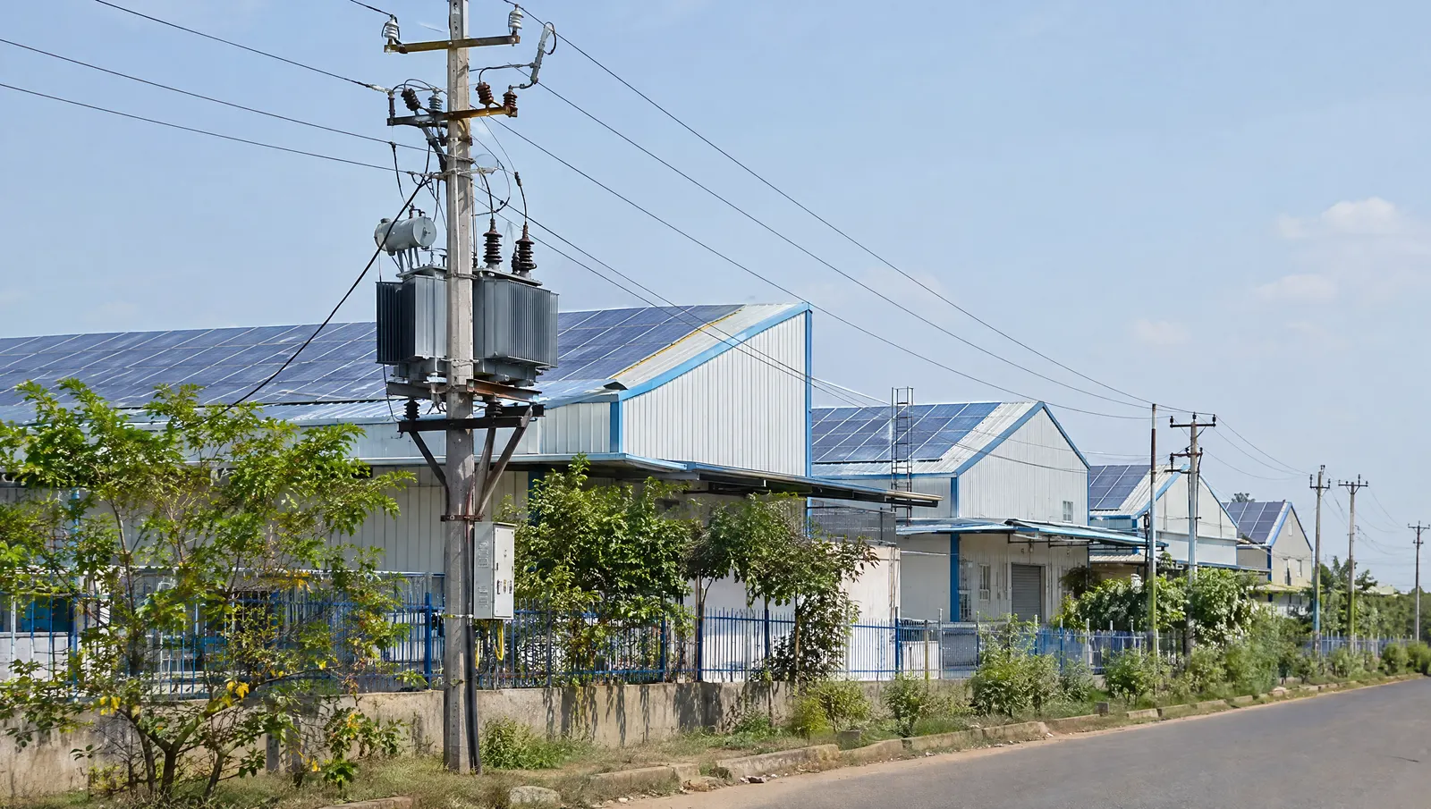 Pole-mounted distribution transformer and LT lines beside Indian industrial sheds with rooftop solar, where surplus units flow back to the grid.